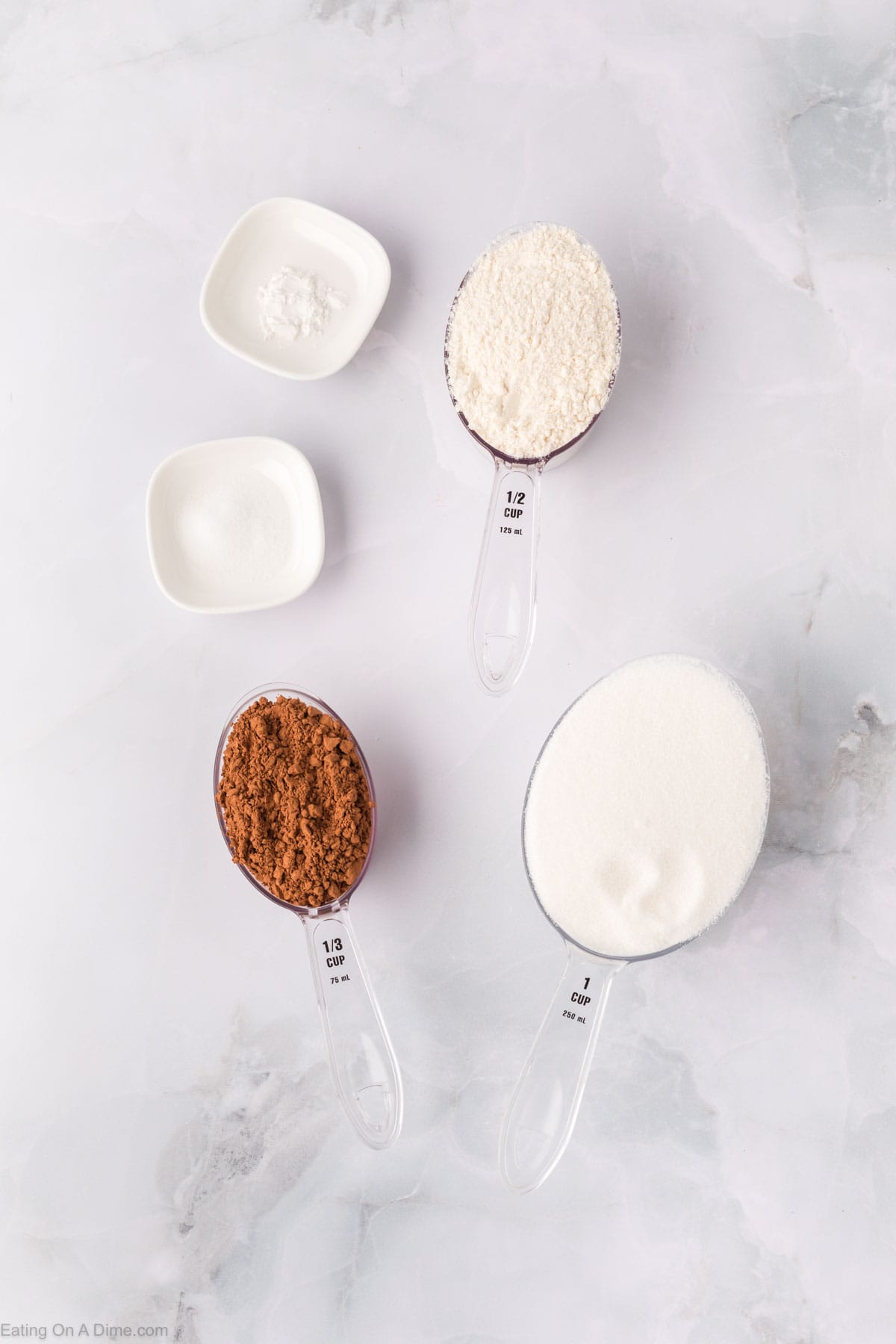 Homemade brownie mix ingredients are beautifully displayed in measuring cups and bowls on a marble surface, featuring flour, brown sugar, white sugar, and a smidgen of baking powder.