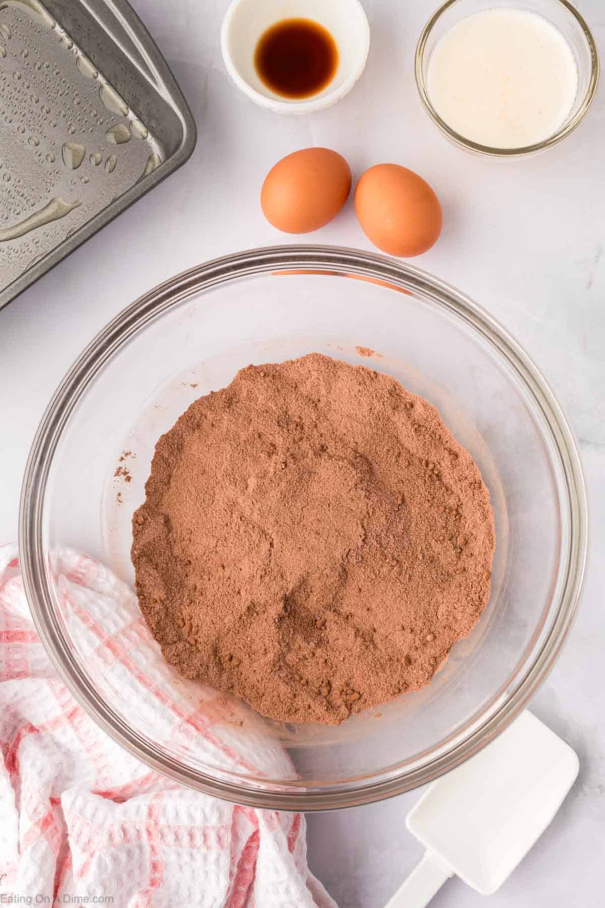 A glass bowl filled with cocoa powder sits on a marble countertop, part of the homemade brownie mix. Nearby are two eggs, a small bowl of vanilla extract, and another bowl with milk. A kitchen towel, a white spatula, and a greased baking pan are also visible.