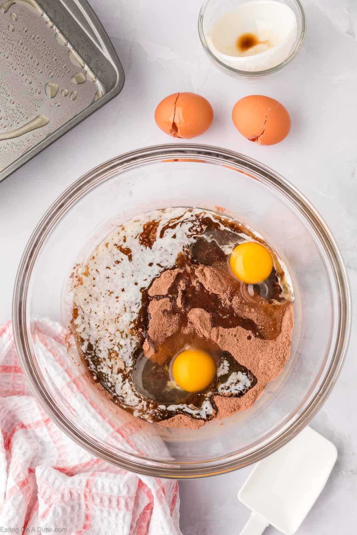 A glass bowl holds ingredients for homemade brownie mix, including cocoa powder, two cracked eggs, and liquid ingredients. Nearby are cracked eggshells, a small bowl with vanilla, a greased baking tray, and a red-and-white kitchen towel.