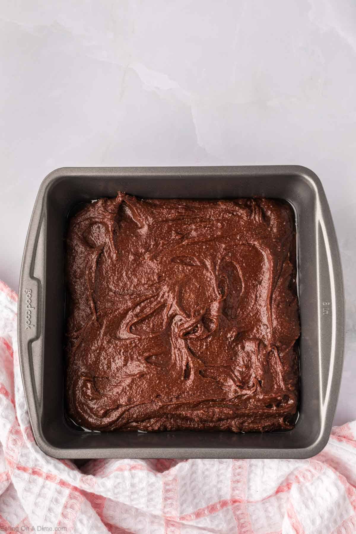 A square baking pan filled with homemade brownie mix, ready to be baked into a delicious chocolate cake. The pan sits on a light gray surface, with a pink and white checkered cloth beside it, adding a cozy touch to the scene.