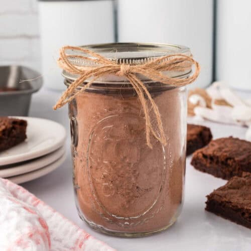 A glass jar filled with homemade brownie mix, tied with a rustic twine bow, sits invitingly on the kitchen counter. In the background, brownie squares and stacked white plates create a cozy baking scene, promising a delightful treat.