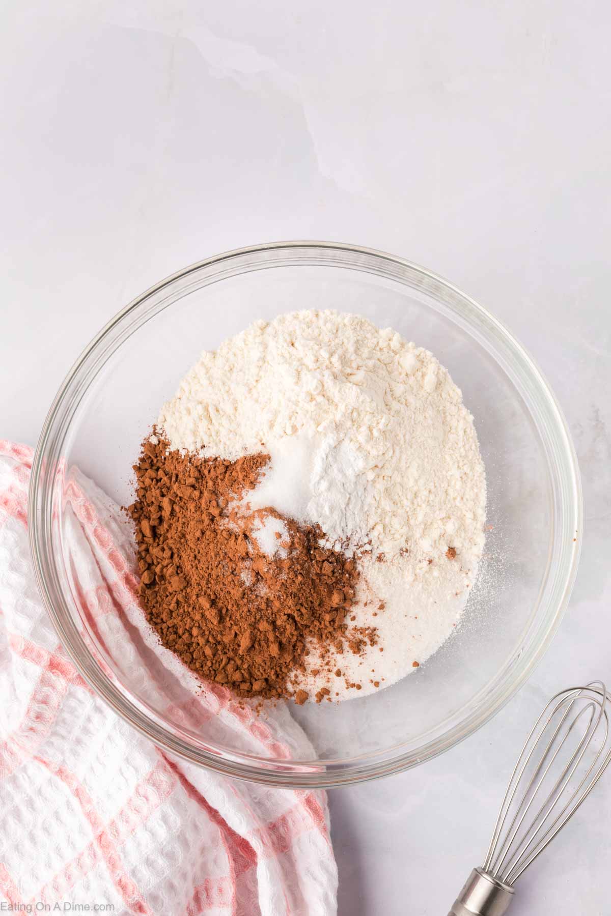 A glass bowl with homemade brownie mix, showcasing flour, cocoa powder, and sugar on a marble surface. A pink and white checkered cloth is beside the bowl, and a whisk is partially visible.