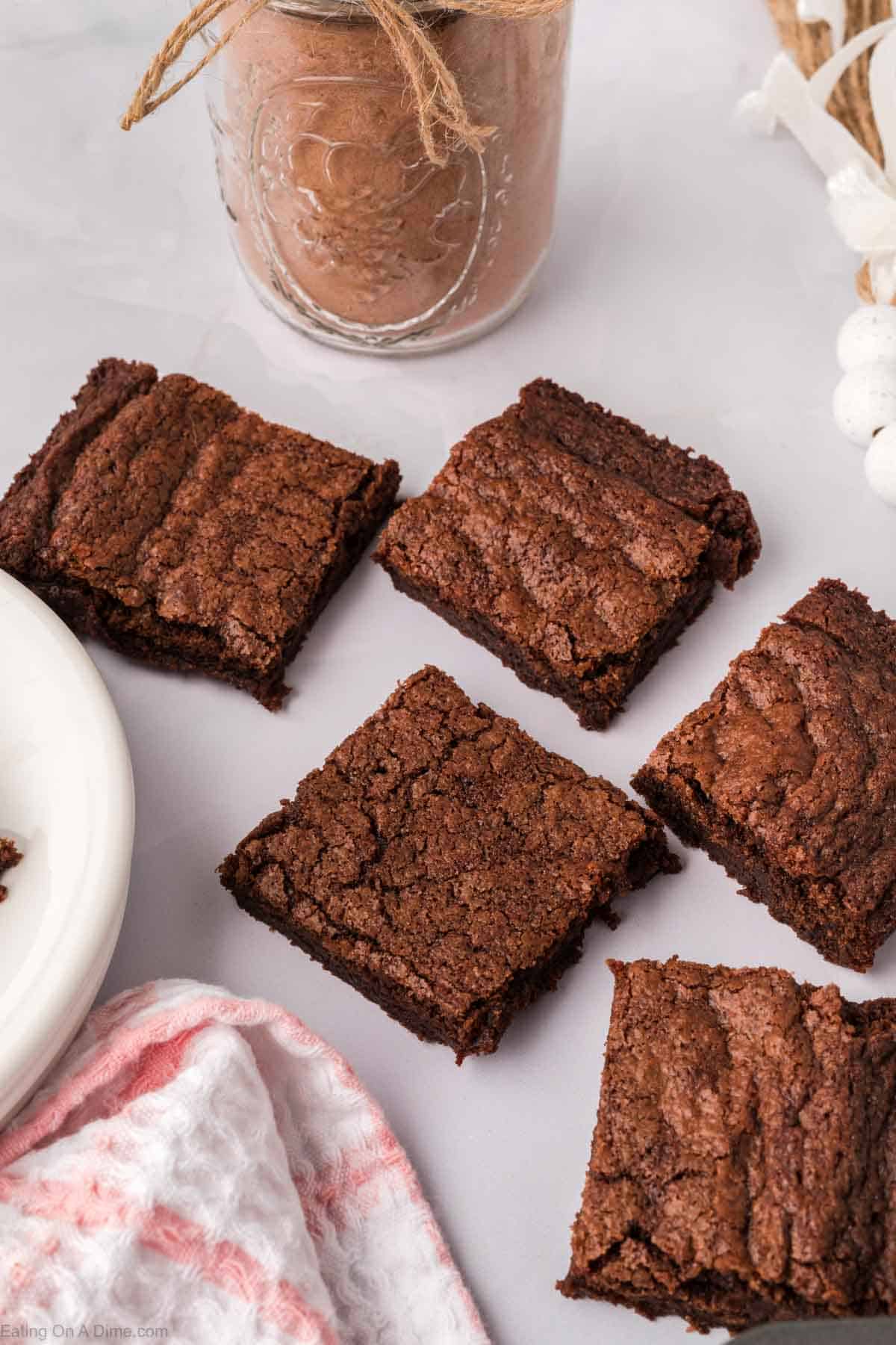 Five chocolate brownies made from a homemade brownie mix rest on a white surface next to a jar with a brown drink tied with twine. A red and white cloth is partially visible on the side.