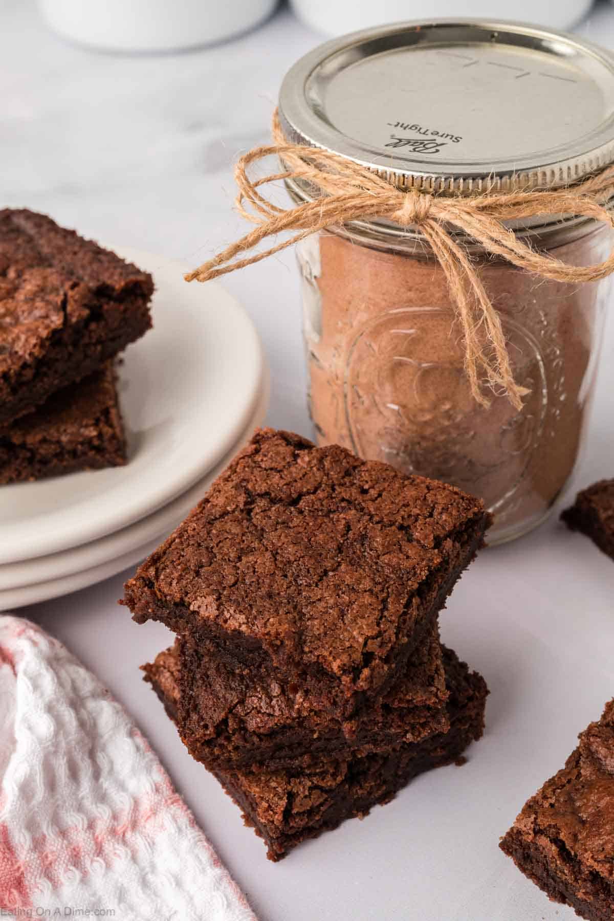 A stack of chocolate brownies sits next to a jar filled with homemade brownie mix, tied with twine. More brownies are on white plates in the background, and a pink and white cloth is nearby on a marble surface.