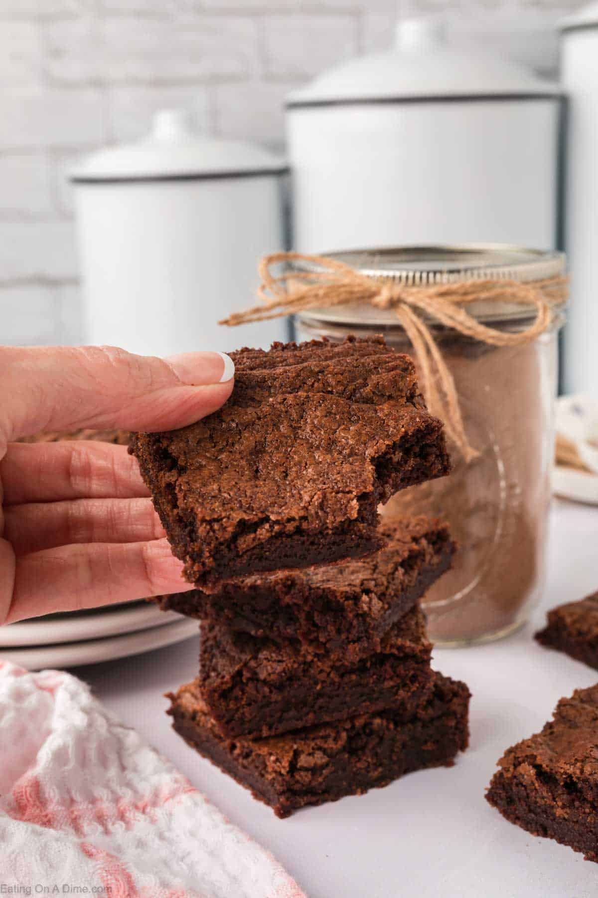 A hand holding a piece of a stacked brownie with a bite taken out, crafted from homemade brownie mix. Several brownies are stacked on a white table next to a jar tied with twine, while two white containers grace the background.