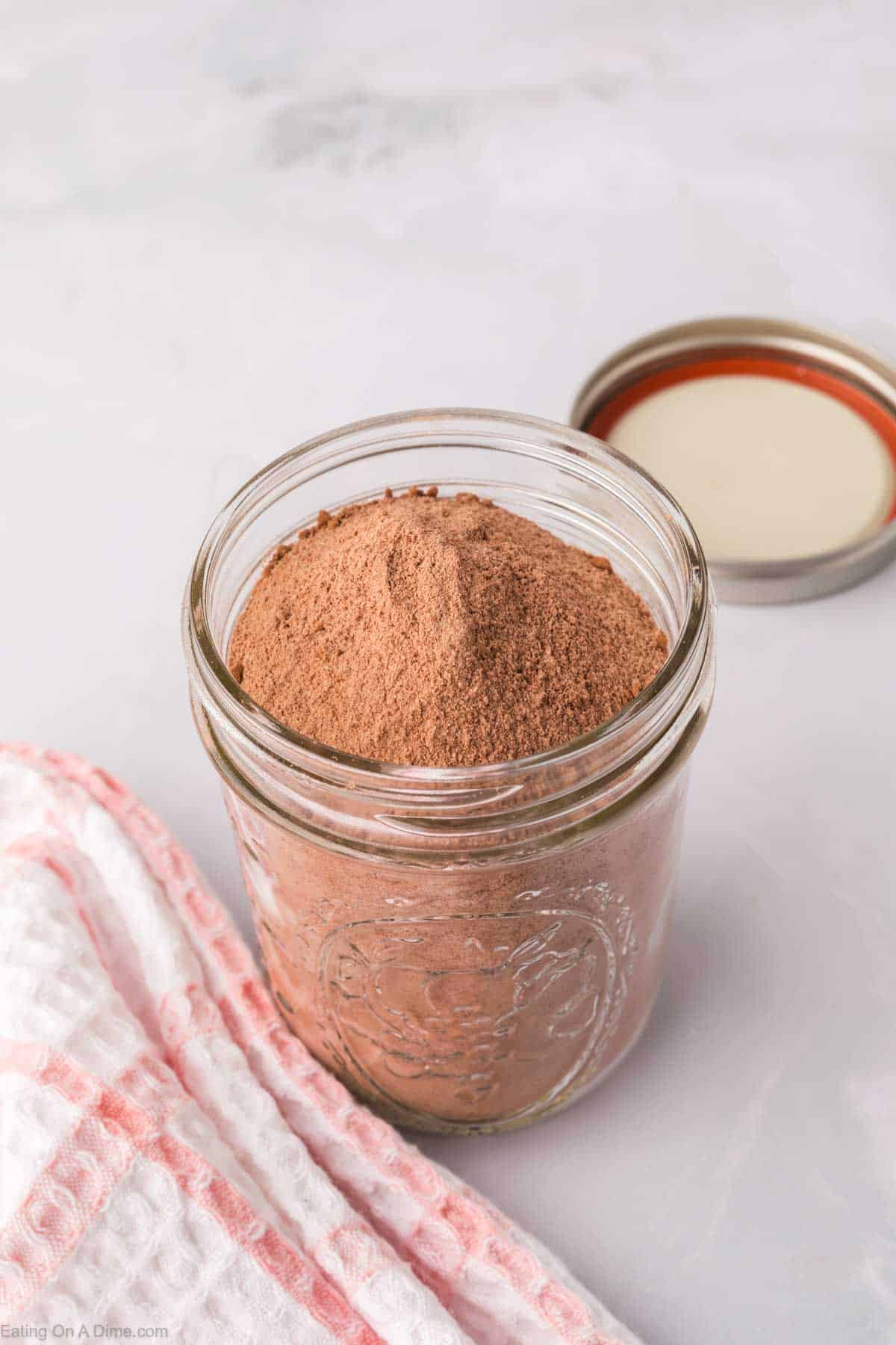 A glass jar filled with homemade brownie mix sits on a light gray surface, its metal lid resting nearby. A textured cloth with red and white stripes is partially visible in the foreground, adding a cozy touch to this inviting scene.