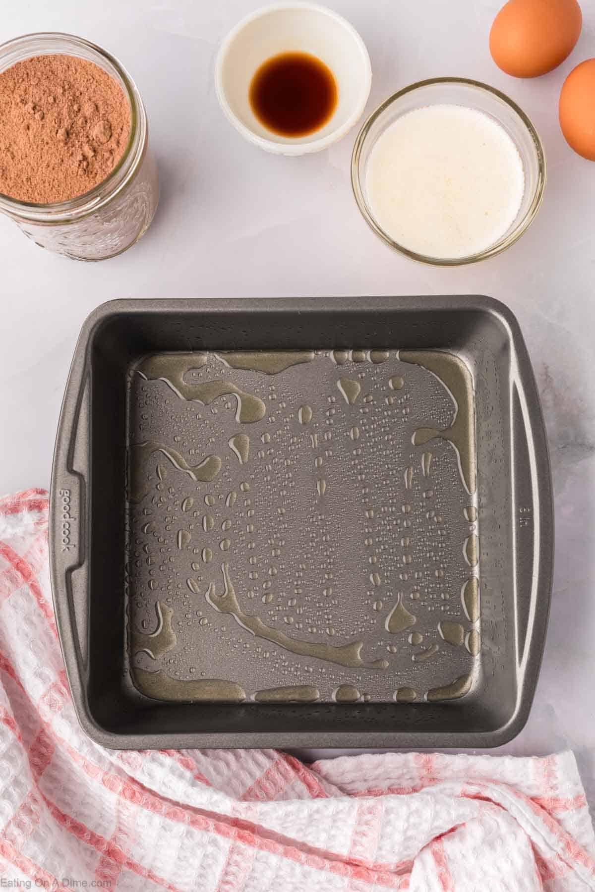 A greased square baking pan awaits on a marble surface. Surrounding it is a delightful array of ingredients for whipping up homemade brownie mix, including a jar of cocoa powder, a bowl of vanilla extract, a bowl of milk, and two eggs. A pink and white towel is nearby.