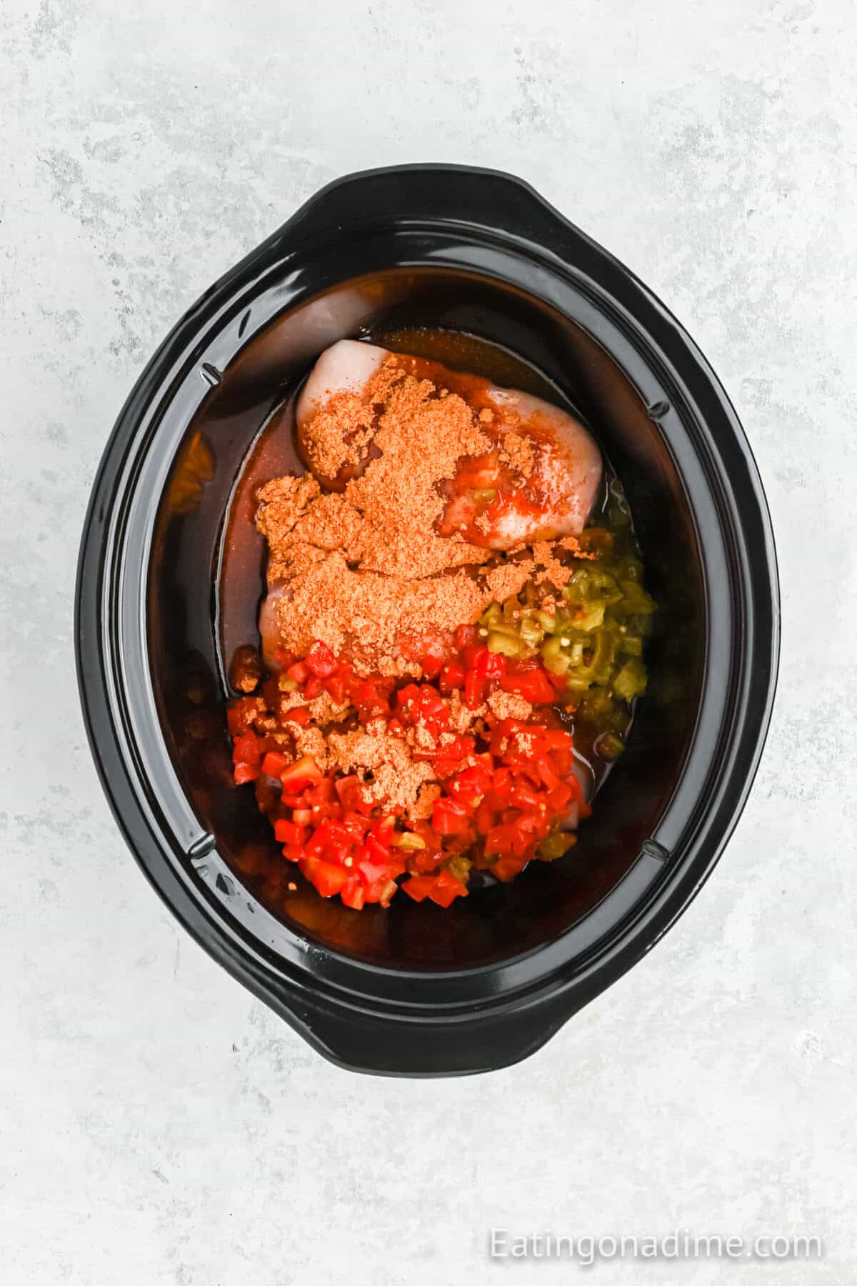 A black crockpot containing raw chicken breasts topped with diced red and green bell peppers, crushed tomatoes, and seasonings for delicious Queso Chicken Tacos. The ingredients are uncooked and ready to be transformed into a slow-cooked masterpiece.