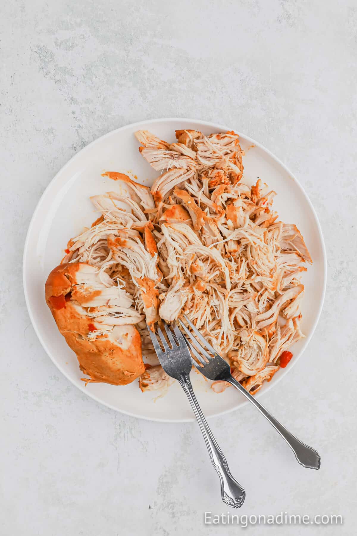 A plate of shredded cooked chicken, perfect for Queso Chicken Tacos, with two forks placed on top, set against a light grey background. The chicken appears tender and moist, ready for serving or further preparation.