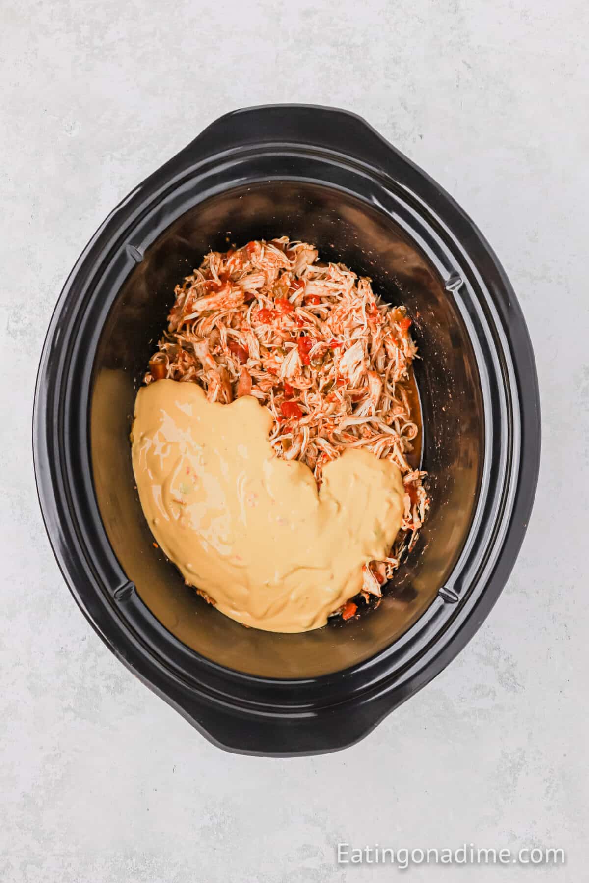 A slow cooker filled with queso chicken tacos features shredded chicken mixed with creamy melted cheese sauce, sprinkled with red spices, and placed against a light gray background.