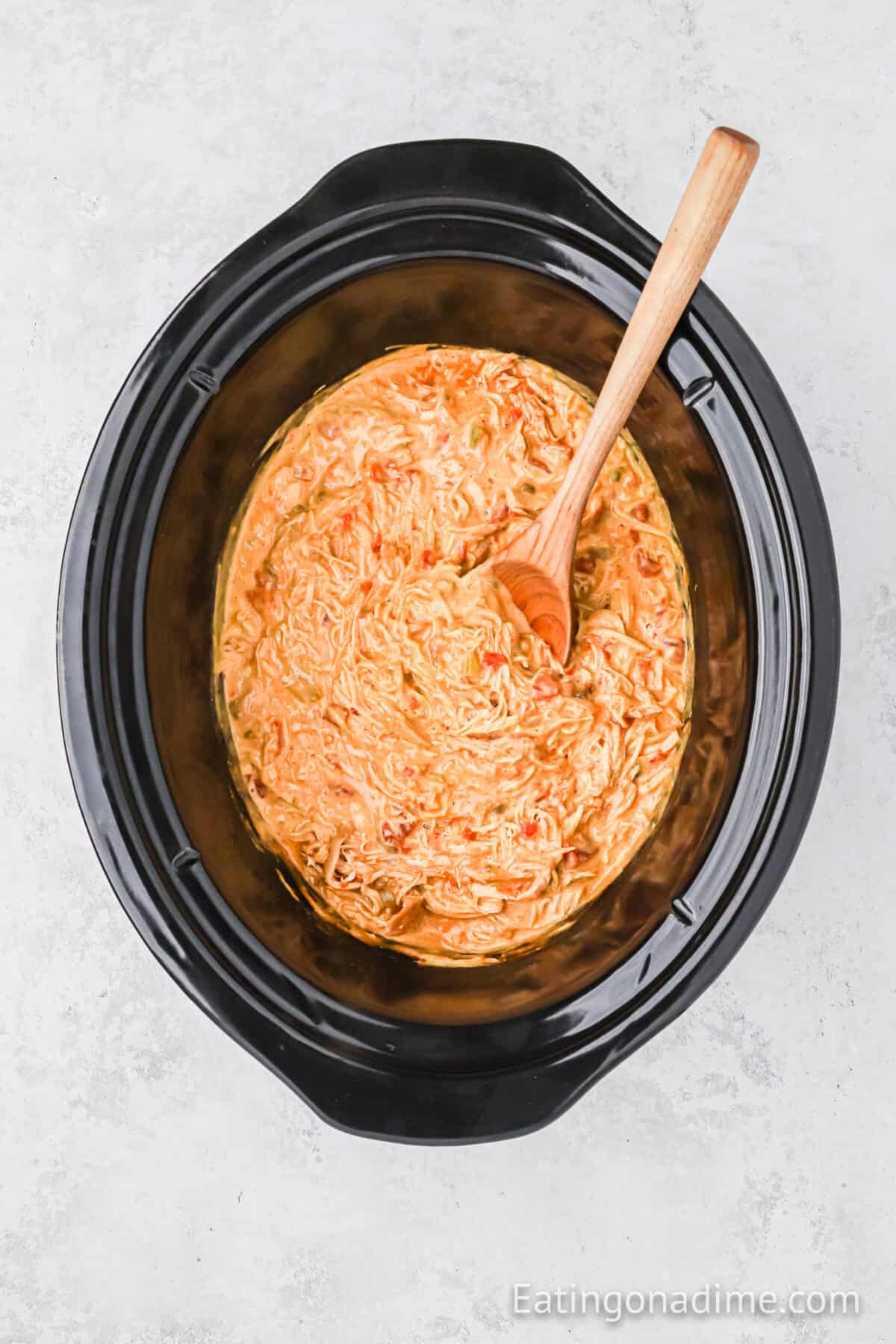 A black slow cooker filled with creamy buffalo chicken dip, featuring shredded chicken and a tangy orange sauce perfect for queso chicken tacos. A wooden spoon rests in the dip, ready for serving.