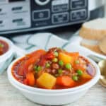 A bowl of hearty vegetable stew with chunks of potato, carrots, peas, and ground beef in a rich tomato sauce. The dish sits on a table with sliced bread in the background and a crock pot subtly visible behind for that perfect slow-cooked touch.