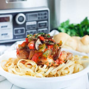 A dish of pasta topped with a blend of slow cooker chicken cacciatore, mushrooms, and peppers in a savory sauce, garnished with herbs. Fresh parsley is blurred in the background. Bread rolls are visible on the side.