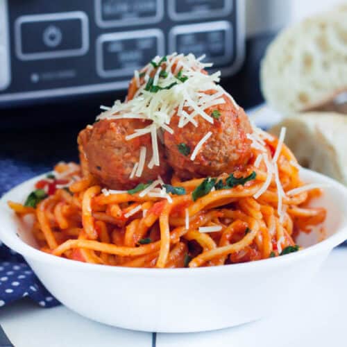 A white bowl brimming with crockpot spaghetti topped with rich tomato sauce, two meatballs, grated cheese, and parsley. A piece of bread is visible in the background, completing this perfect recipe scene.