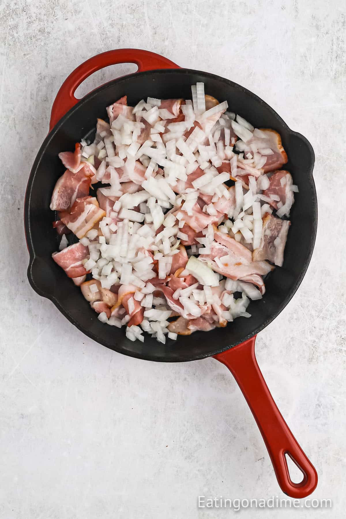 A red-handled skillet contains chopped bacon and diced onions, ready to cook. Beside it sits a slow cooker filled with cabbage on a light-colored surface, promising a hearty meal in the making.