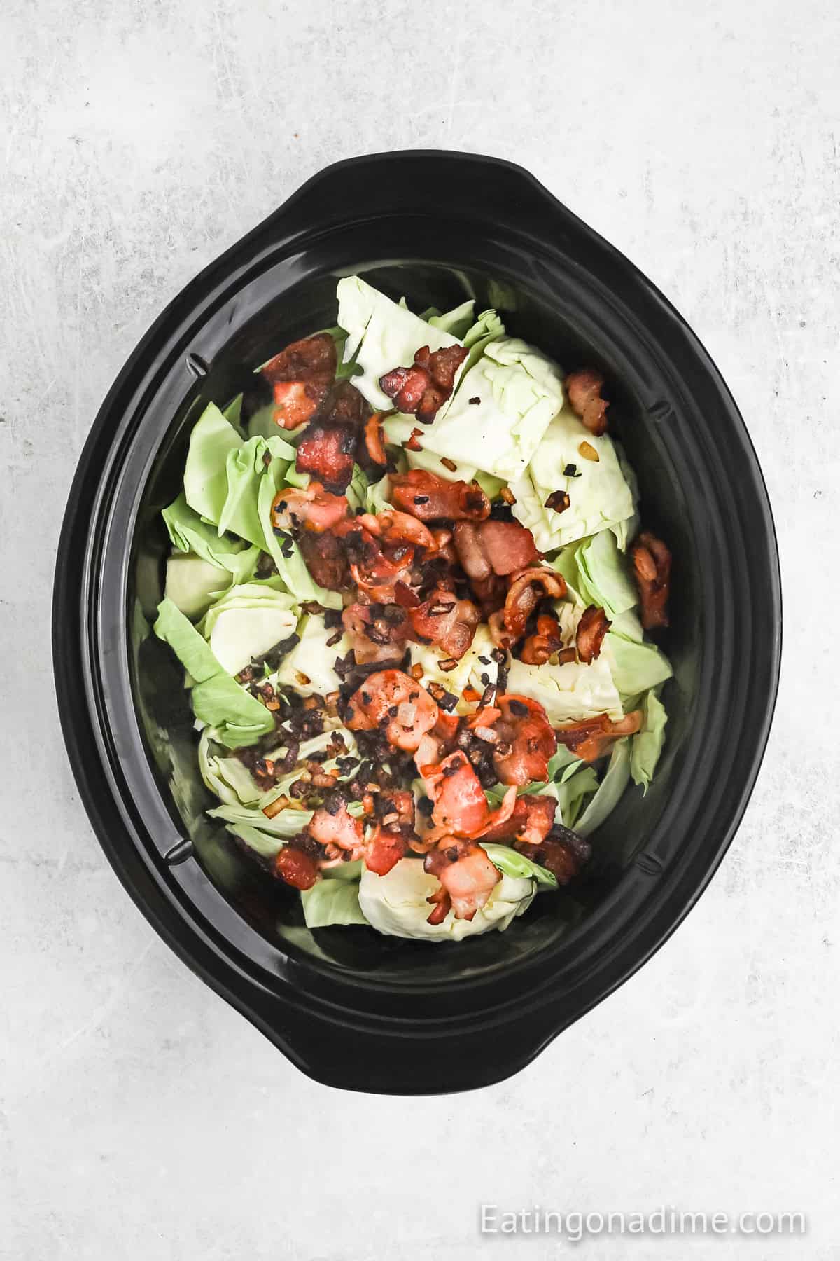 Slow Cooker Cabbage with bacon pieces sits invitingly in a black slow cooker against a light textured background.