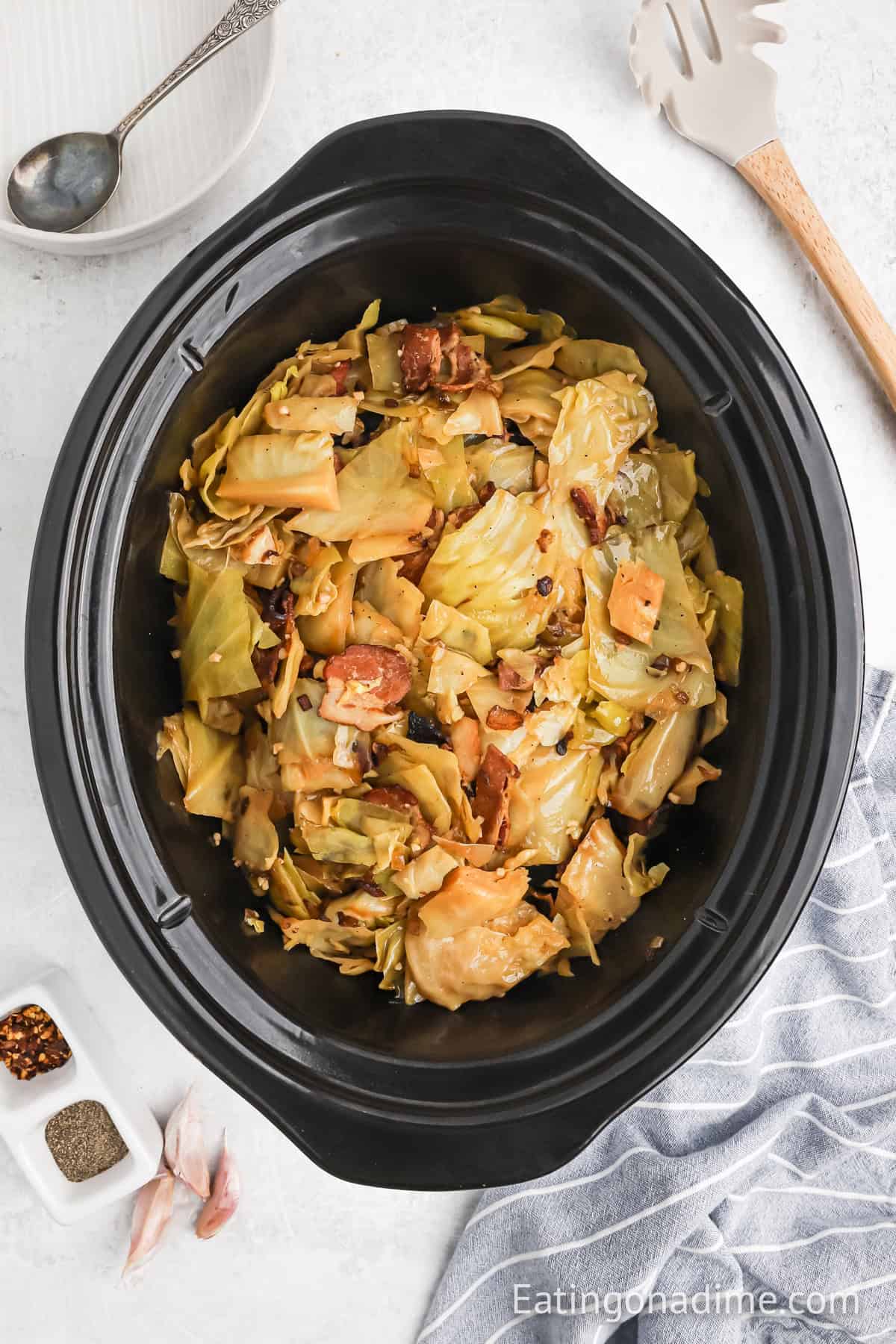 Slow Cooker Cabbage with bacon pieces simmering in a black crockpot. A striped cloth sits beside the pot, alongside a spoon and white dish in the background. Small bowls of spices are also nearby, ready to enhance the hearty flavors of this cozy dish.