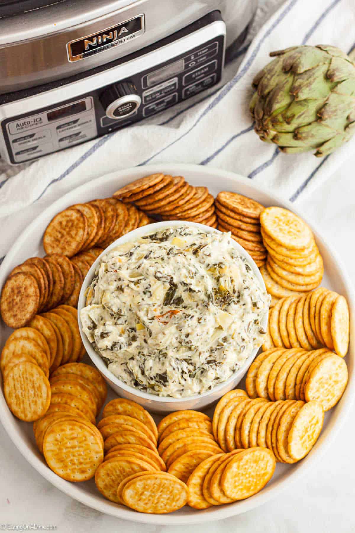 A bowl of creamy Crockpot Spinach Artichoke Dip sits on a white plate, surrounded by a ring of round crackers. A slow cooker and a fresh artichoke rest in the background on a striped cloth, inviting everyone to savor this delicious creation.