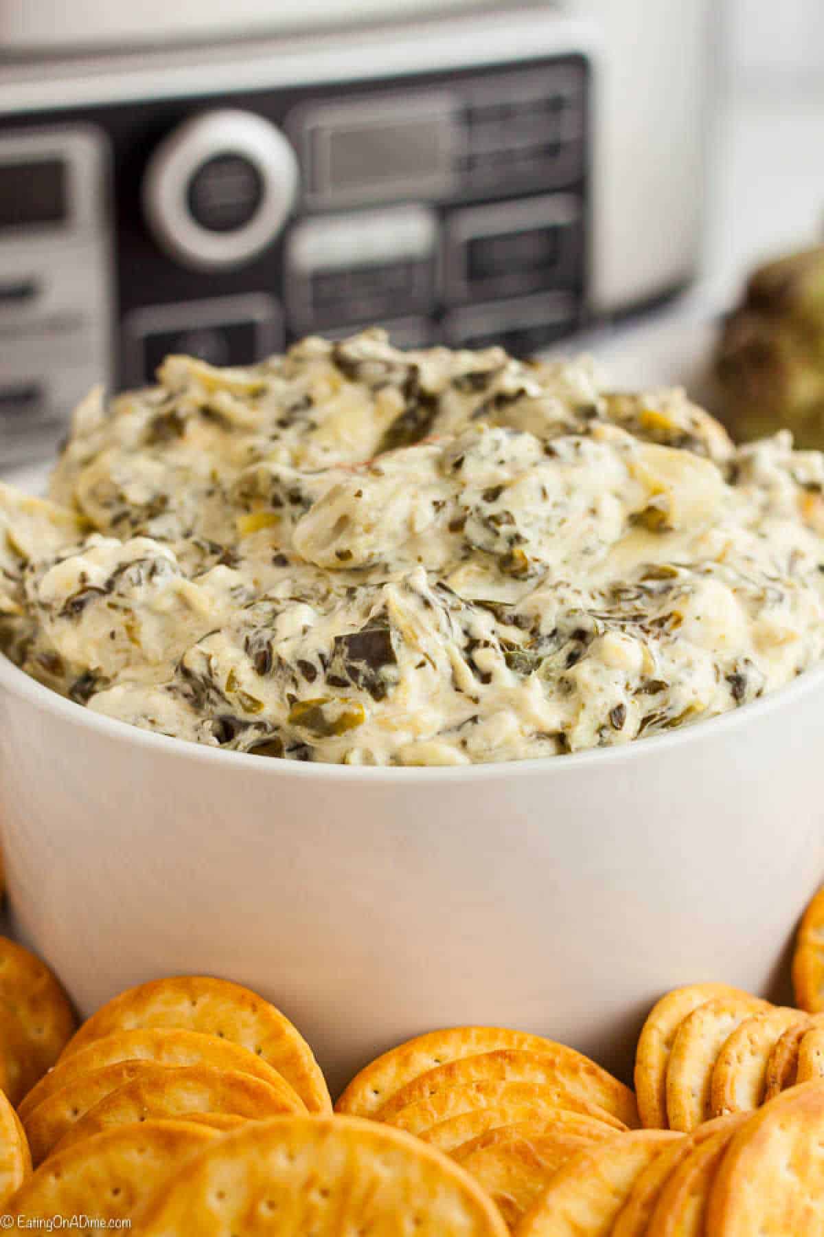 A white bowl filled with creamy Crockpot Spinach Artichoke Dip is surrounded by an arrangement of round crackers, with a slow cooker subtly visible in the background.