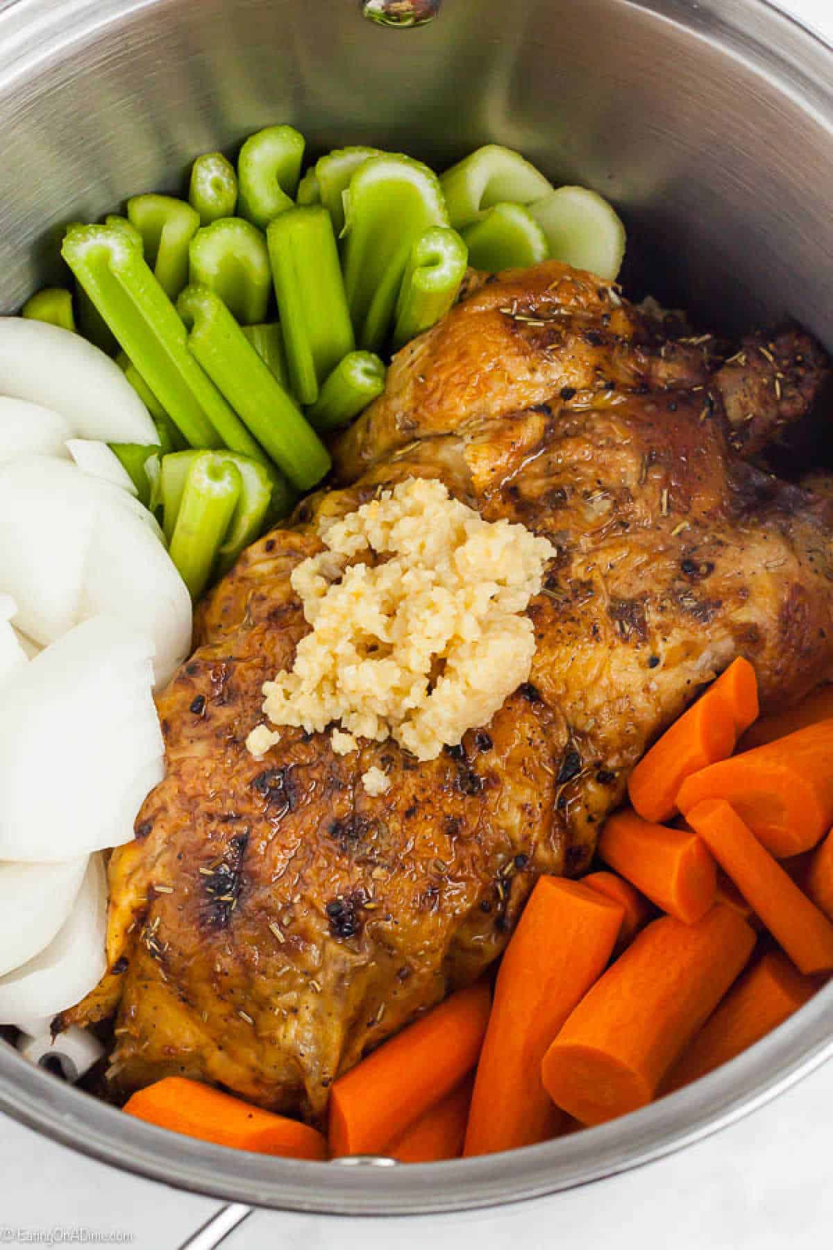 A cooked seasoned roast chicken topped with minced garlic is surrounded by chopped onions, celery, and carrots in a stainless steel pot, perfect for those learning how to make chicken stock.