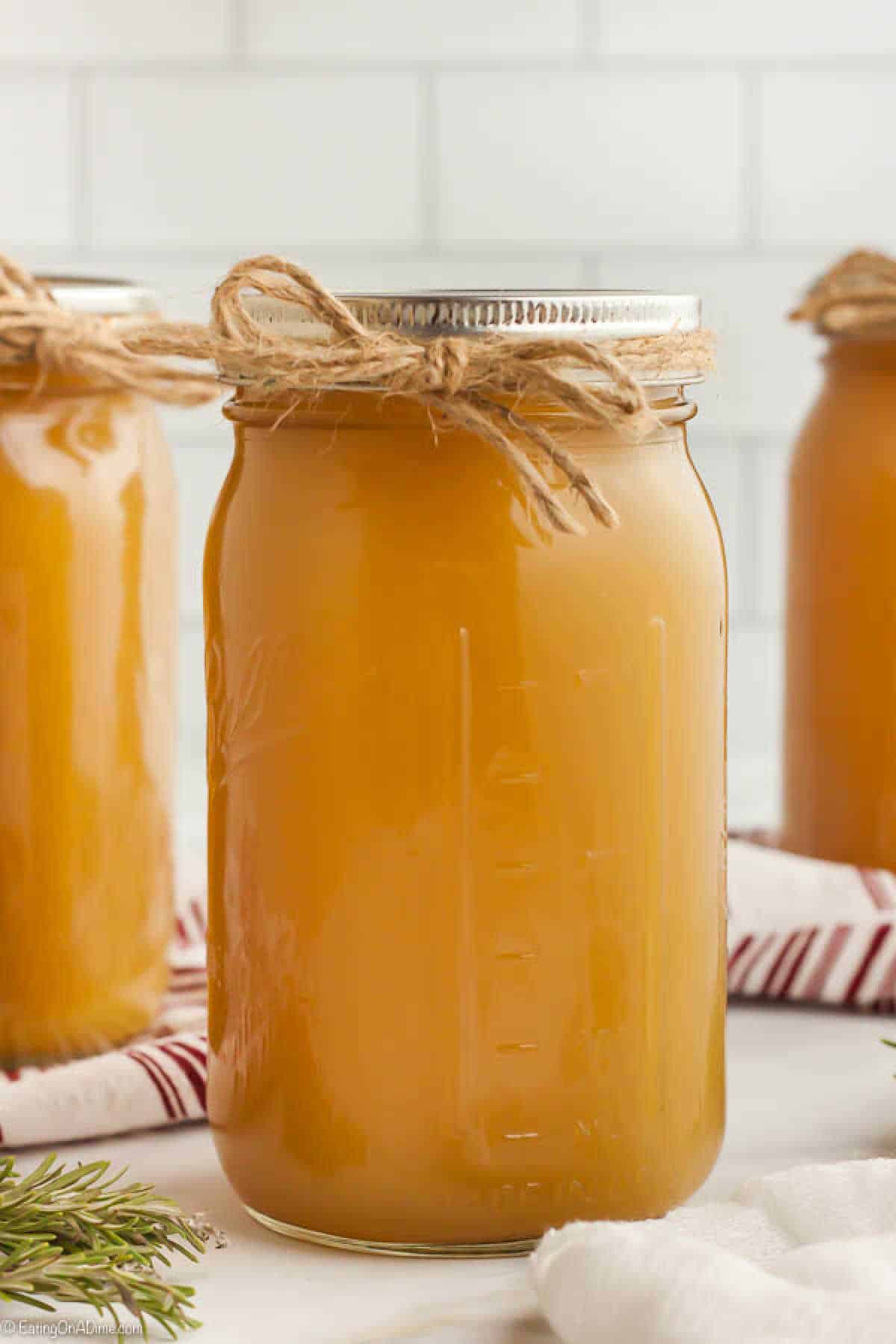 A glass jar filled with a light brown liquid, sealed with a metal lid and tied with a rustic twine bow around the top—an inviting sight for anyone learning how to make chicken stock. Two similar jars are partially visible in the background, set on a white surface with a striped cloth and greenery nearby.