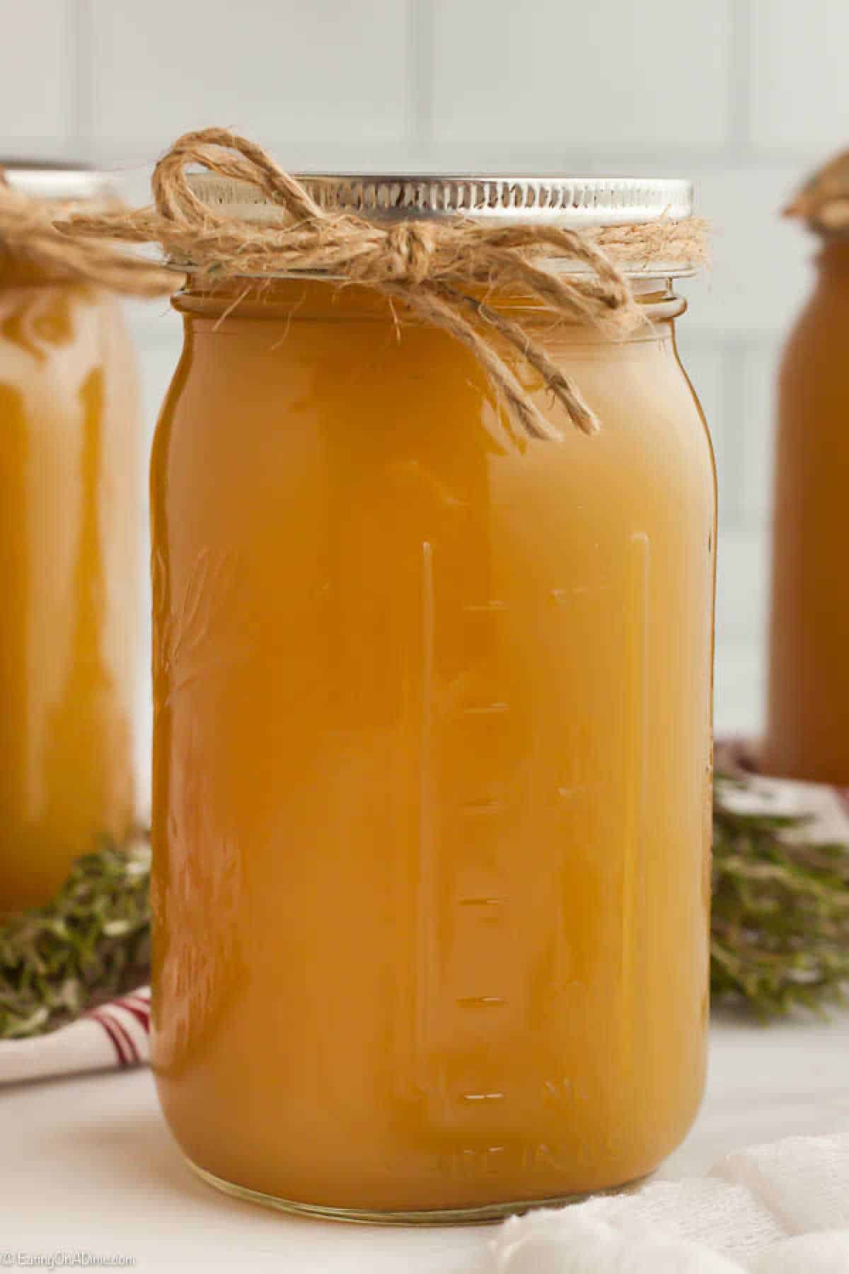 A glass jar filled with golden brown liquid, likely homemade stock, with a metal lid and a jute twine bow tied around the top. The background shows more jars of the same liquid and hints of green herbs on a light-colored surface—a perfect illustration of how to make chicken stock at home.