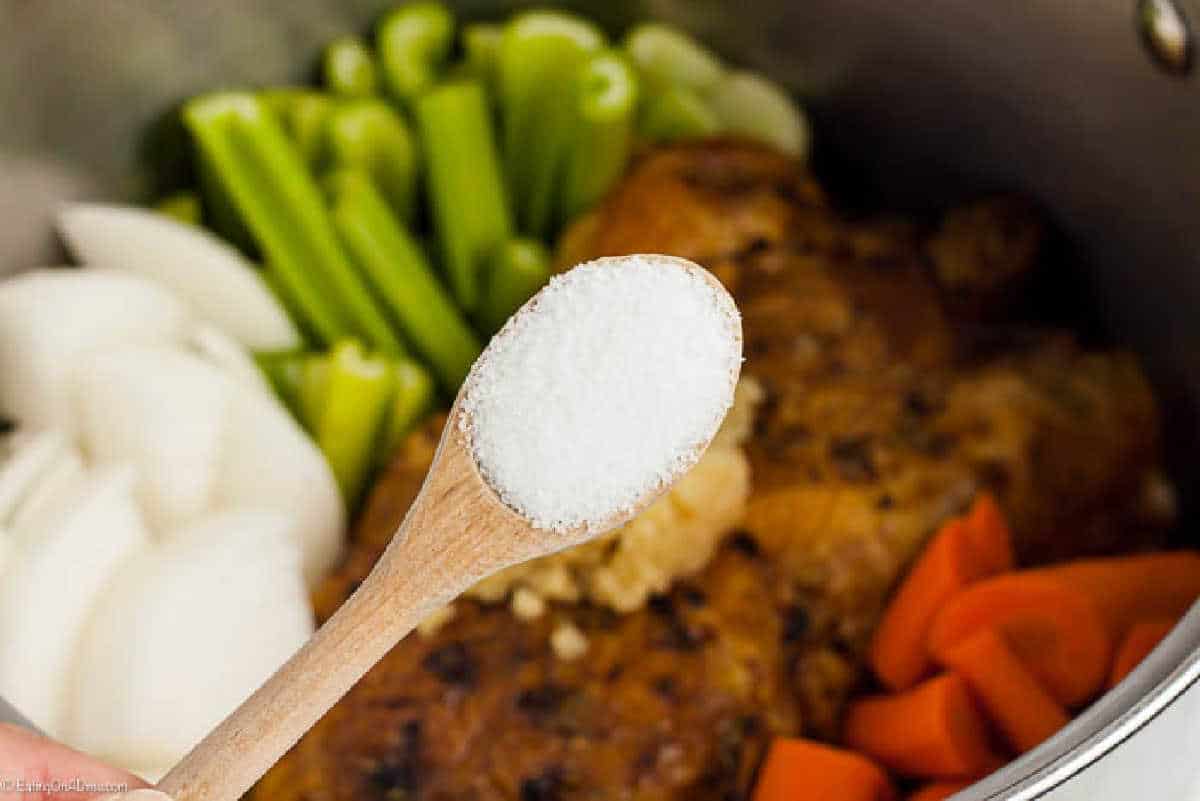 A wooden spoon filled with salt is held over a pot containing cooked chicken, onion wedges, chopped celery, and carrot pieces, ready to be seasoned—perfect steps on how to make chicken stock.