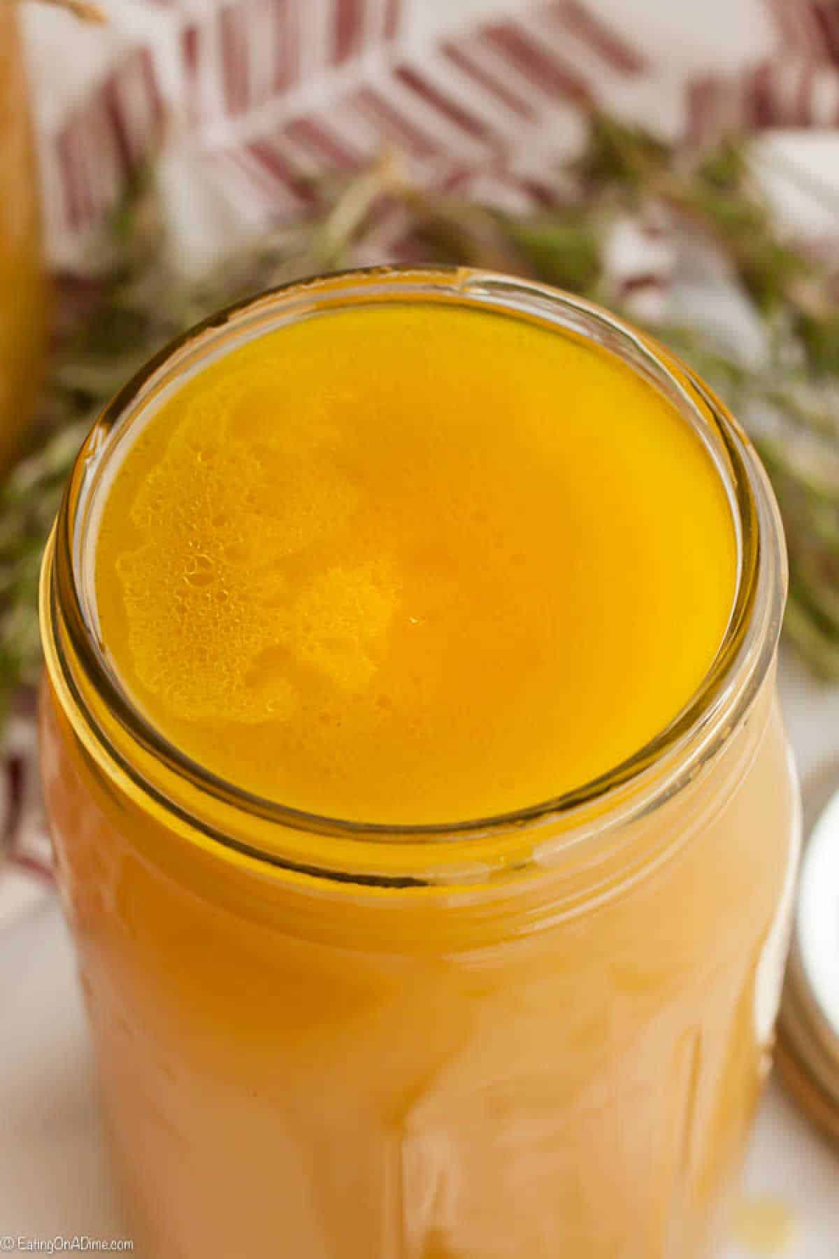 A close-up of a glass jar filled with golden yellow liquid, likely homemade ghee or clarified butter. The surface of the liquid shows some bubbles and foam. In the background, there are sprigs of herbs and a partially visible cloth with a red pattern—perfect ingredients for how to make chicken stock.