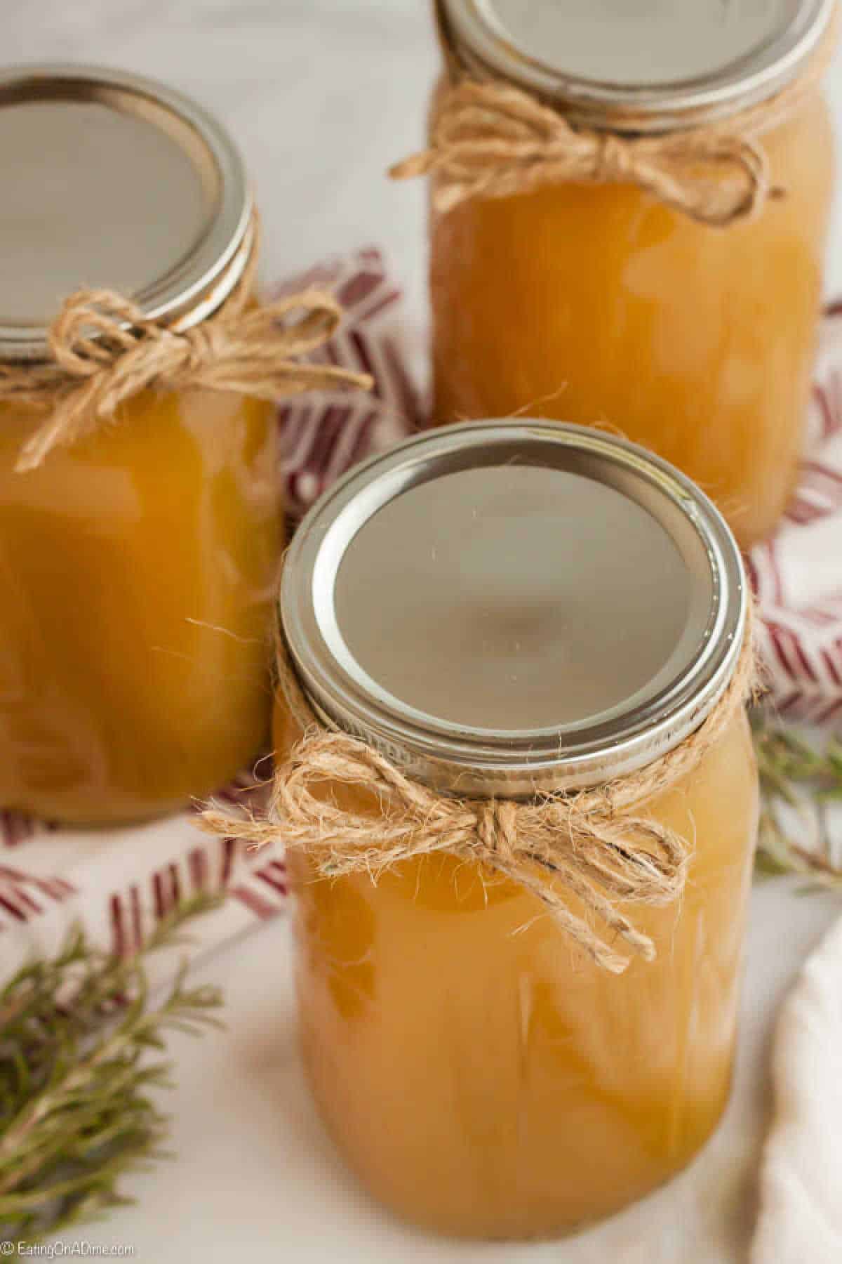 Three glass jars filled with light brown liquid, likely a homemade broth or sauce, are arranged on a white surface. Each jar is sealed with a metallic lid and decorated with a twine bow around the neck. Greenery is partially visible at the bottom left, adding charm to this guide on how to make chicken stock.