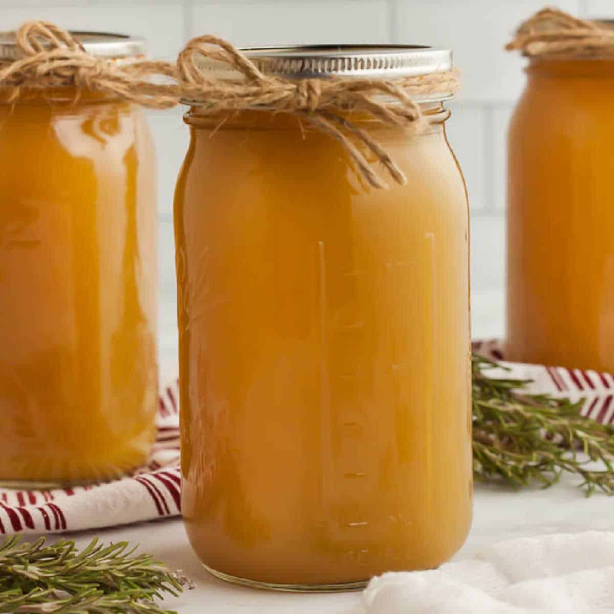 Three jars filled with light brown liquid, likely homemade chicken stock or juice, are sealed with metal lids and a twine bow around the neck. Fresh rosemary sprigs and a red-striped dish towel are placed around the jars on a white countertop.