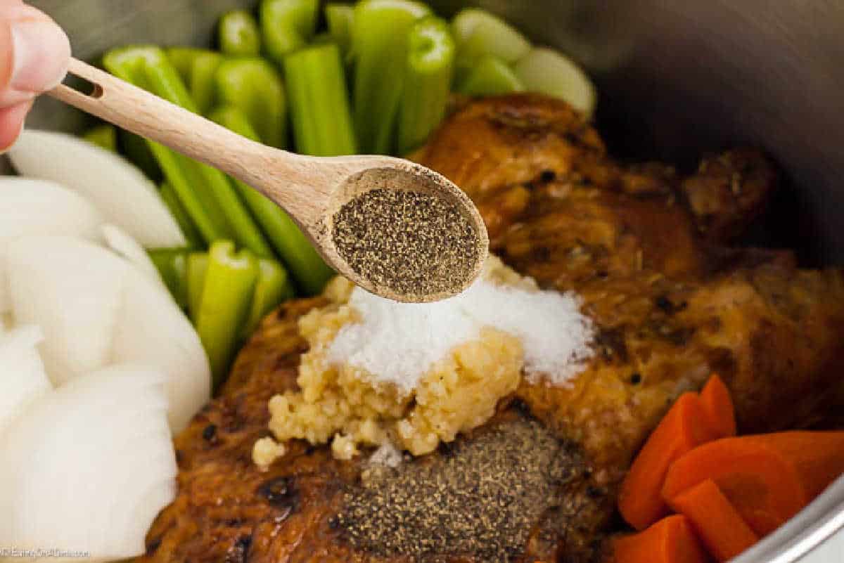 A wooden spoon holding ground black pepper hovers over a cooked chicken surrounded by chopped onions, celery, and carrots in a cooking pot. Salt and minced garlic are also visible on top of the chicken, illustrating how to make chicken stock from scratch.