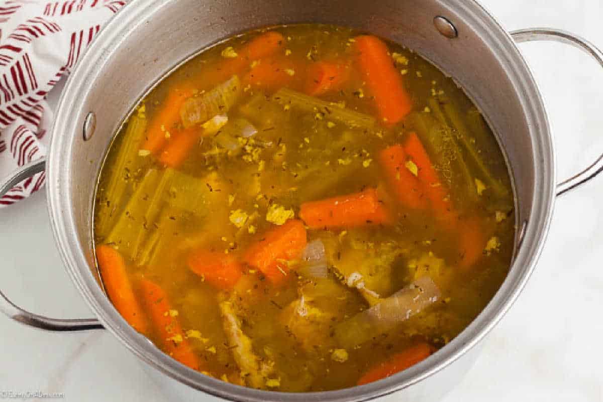 A metal pot filled with homemade chicken broth, featuring chunks of carrots, celery, and chicken pieces. The broth, a perfect example of how to make chicken stock, is seasoned with herbs and has a rich golden color. The pot is placed on a white surface next to a red and white striped cloth.