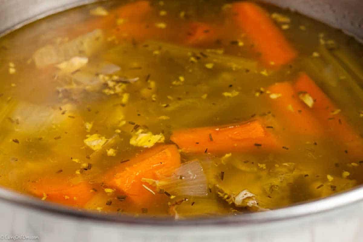 A close-up of a pot filled with steaming chicken soup. The broth, rich from homemade chicken stock, is golden and contains large chunks of orange carrots and pieces of shredded chicken, along with herbs and small bits of onion.