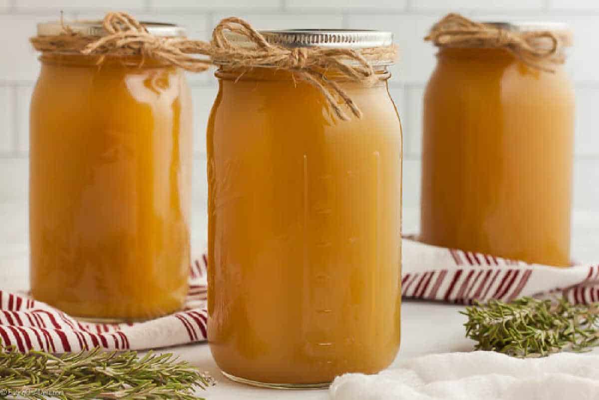 Three mason jars filled with golden liquid, each topped with a metal lid and tied with twine, sit on a white surface. A striped cloth and sprigs of rosemary add charm. It's the perfect setup for learning how to make chicken stock against a white tiled backdrop.