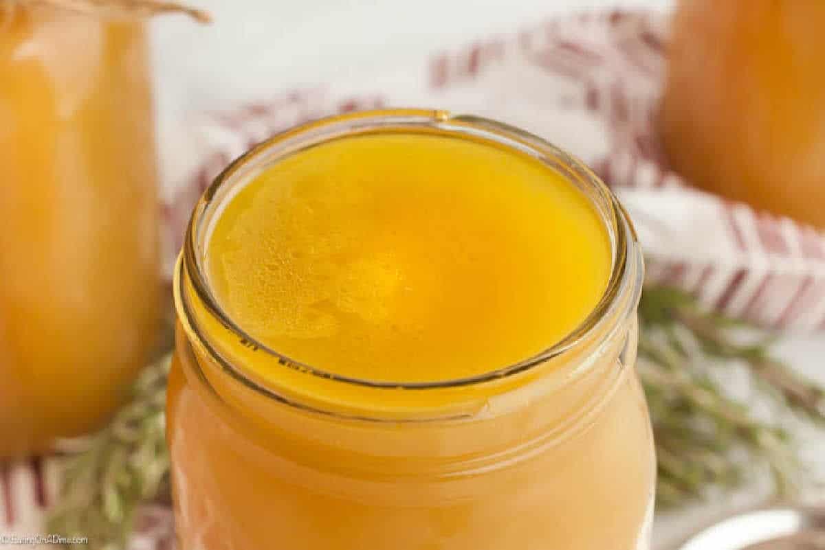 A close-up of the top of an open glass jar filled with golden yellow ghee. The smooth surface of the ghee glistens under light, like a secret ingredient for those learning how to make chicken stock. The background features a blurred red and white patterned cloth and some green foliage.