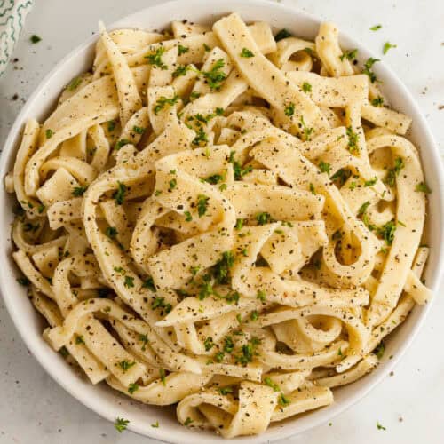 A bowl of homemade egg noodles topped with creamy white sauce, garnished with black pepper and fresh parsley.