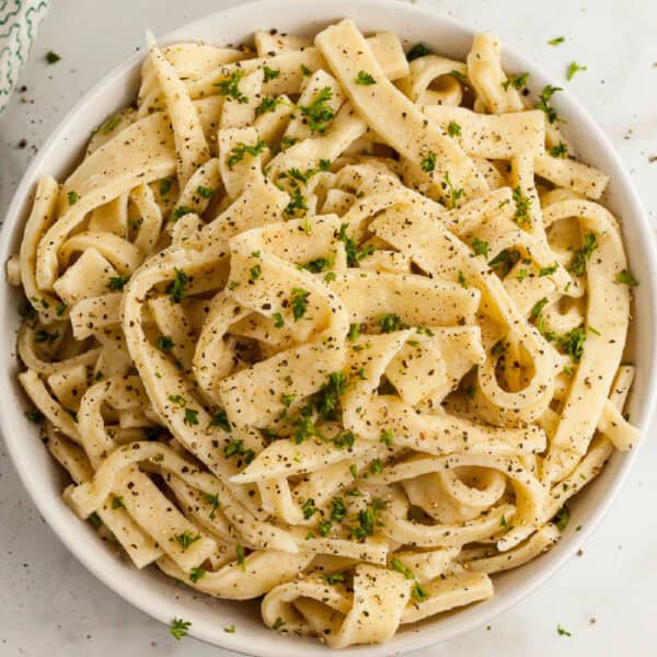 A bowl of homemade egg noodles topped with creamy white sauce, garnished with black pepper and fresh parsley.