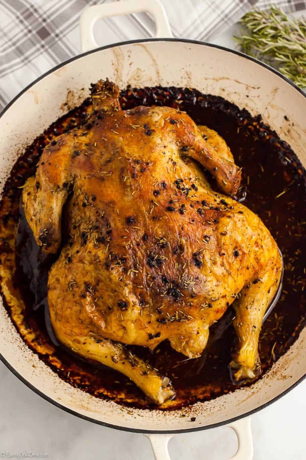 A whole roasted chicken with crispy, golden skin sits in a round white pan, showcasing the perfect "how to roast a chicken" technique. Surrounded by a rich, dark sauce and garnished with herbs, it's framed by a striped cloth and rosemary sprigs in the background.