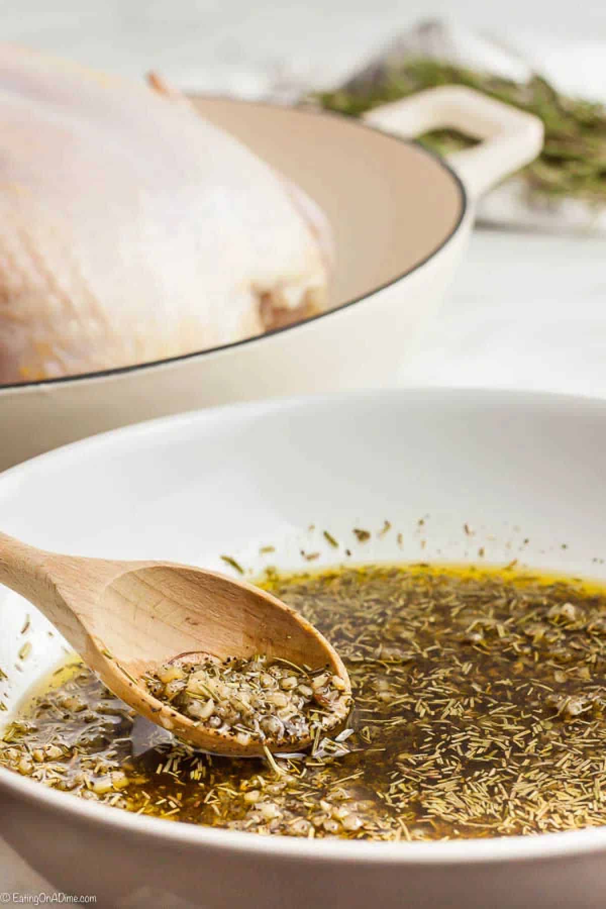 A wooden spoon stirs a herb and garlic marinade in a white bowl, with a raw turkey in a pot nearby, offering hints on how to roast a chicken. The mixture of herbs, garlic, and oil suggests flavorful preparation for roasting.