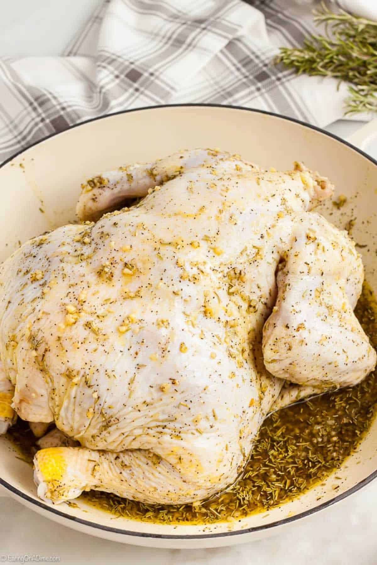 A whole raw chicken seasoned with herbs and spices sits in a large white pot, inviting you to explore how to roast a chicken perfectly. A beige plaid cloth and sprigs of fresh herbs set the scene, suggesting preparation for cooking.