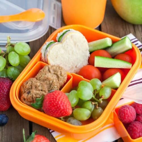 A bright orange lunchbox, perfect for easy lunch ideas for kids, contains a heart-shaped sandwich, cucumber sticks, cherry tomatoes, green grapes, strawberries, and two cookies. It is accompanied by an orange drink container. Additional fruits are scattered nearby on a striped cloth.