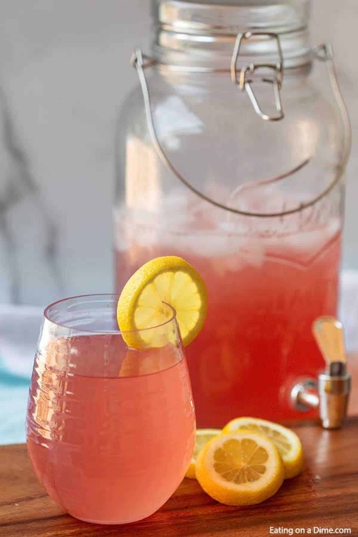 A refreshing glass of pink lemonade, showcasing a zesty lemon slice on the rim, sits elegantly on a wooden surface. Behind it, a large glass dispenser brimming with ice-cold pink lemonade and equipped with a sturdy metal spigot and handle offers easy access to your favorite pink lemonade recipe.