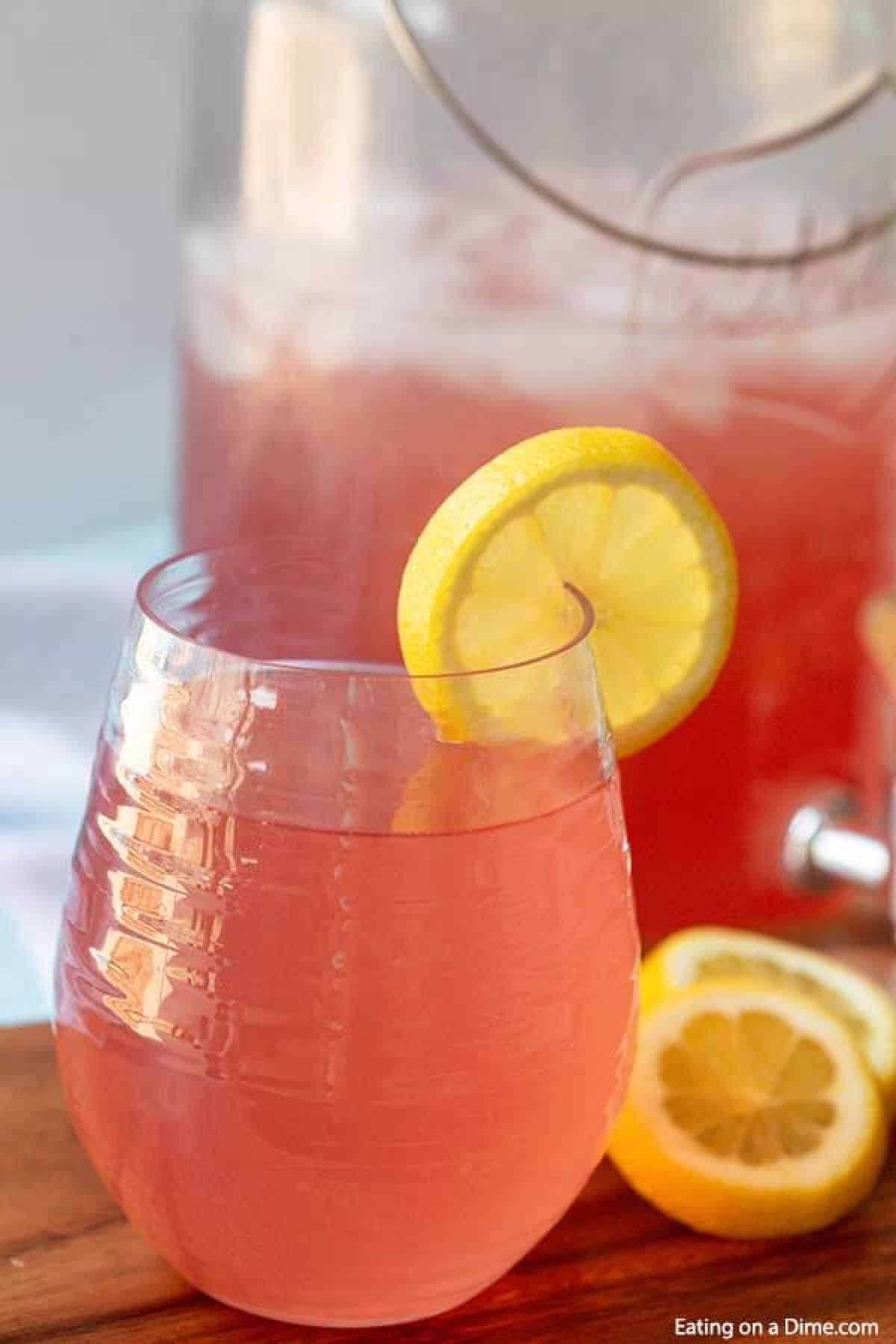 A glass of pink lemonade with a lemon slice garnish sits on a wooden surface, completing this simple yet refreshing pink lemonade recipe. In the background, a pitcher filled with ice and more pink lemonade awaits while a few lemon slices are scattered nearby.