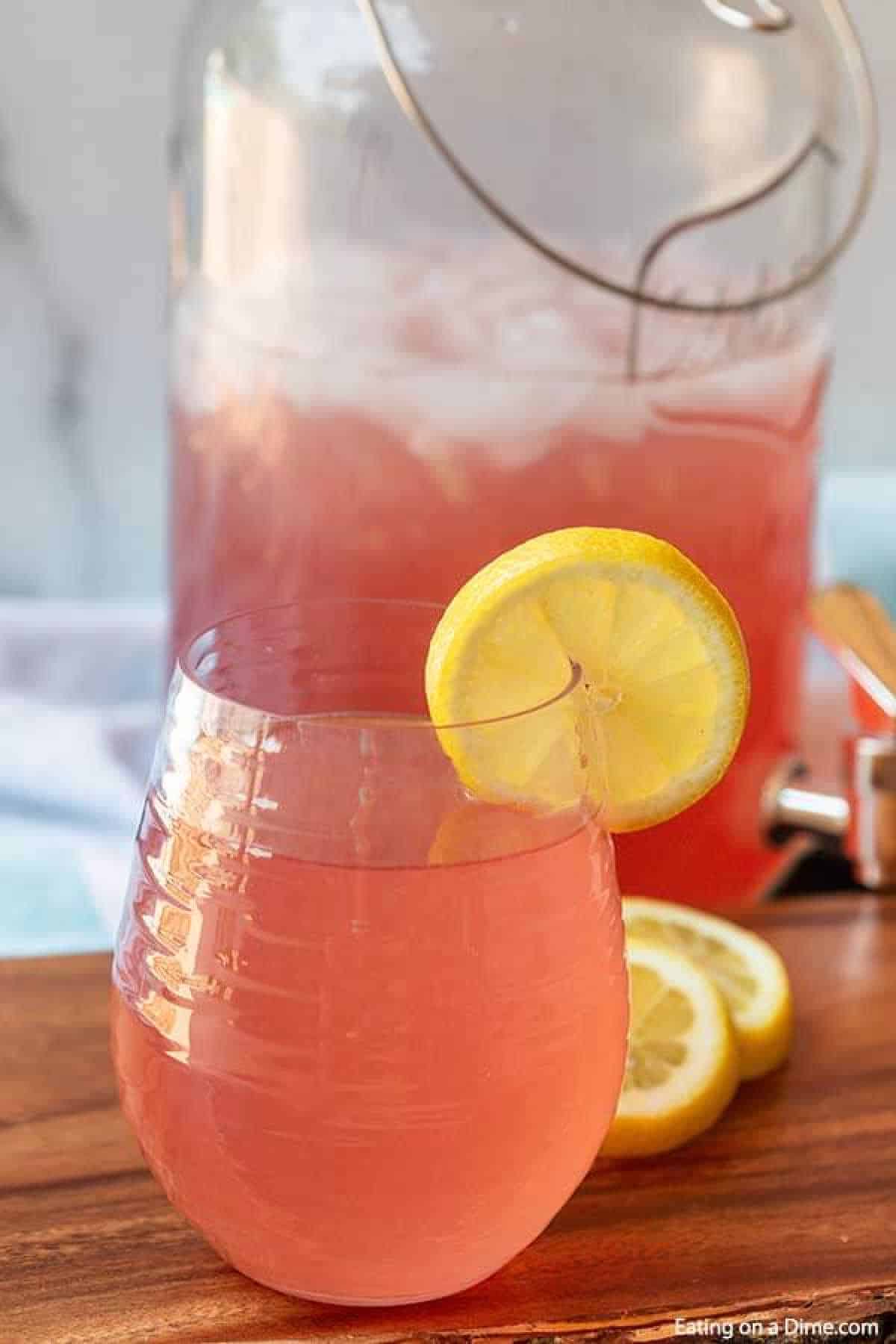 A glass of refreshing pink lemonade with a lemon slice on the rim sits on a wooden surface. Behind it, a large pitcher filled with more pink lemonade and ice awaits, hinting at a delightful pink lemonade recipe. Nearby, slices of lemon add a zesty touch.