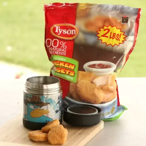 A packet of Tyson chicken nuggets, labeled "2 lbs" and "100% natural ingredients," is placed on a table. Nearby, an insulated container with a cartoon character on it holds several chicken nuggets, with the lid set beside it—perfect easy lunch ideas for kids.