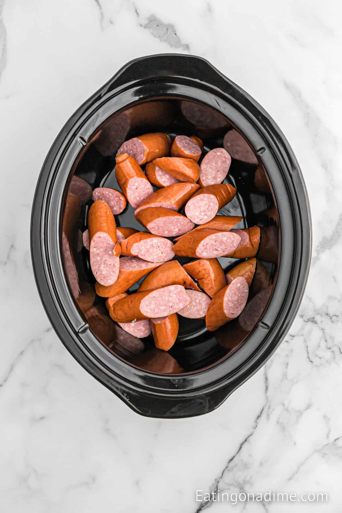 A black slow cooker on a marble surface contains sliced sausages with visible pink interiors, perfect for making Slow Cooker Kielbasa and Sauerkraut. The text "Eatingonadime.com" is visible in the lower right corner of the image.