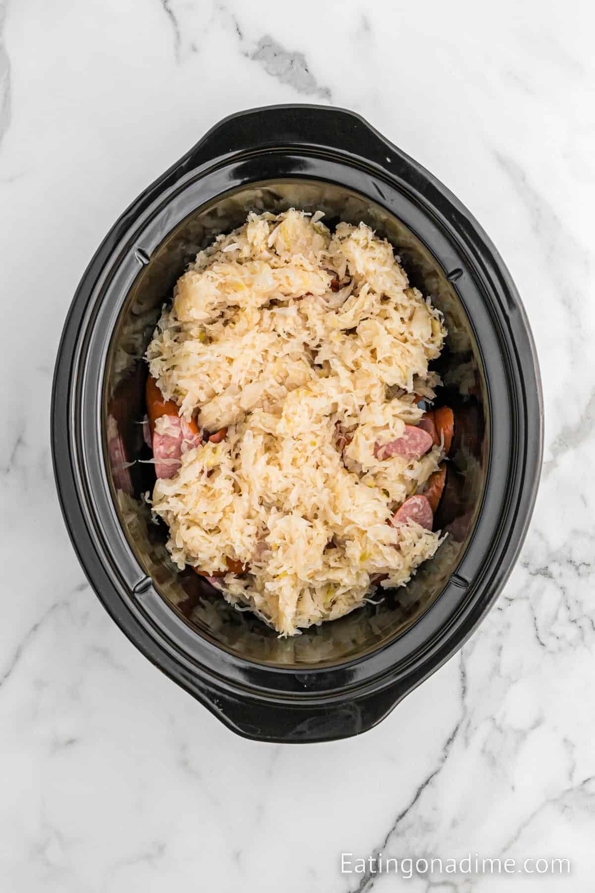 A slow cooker filled with Slow Cooker Kielbasa and Sauerkraut, along with chopped sausages or hot dogs and shredded meat, sits on a marbled countertop. The ingredients appear cooked and well-mixed within the black slow cooker pot. The website "Eatingonadime.com" is visible in the corner.