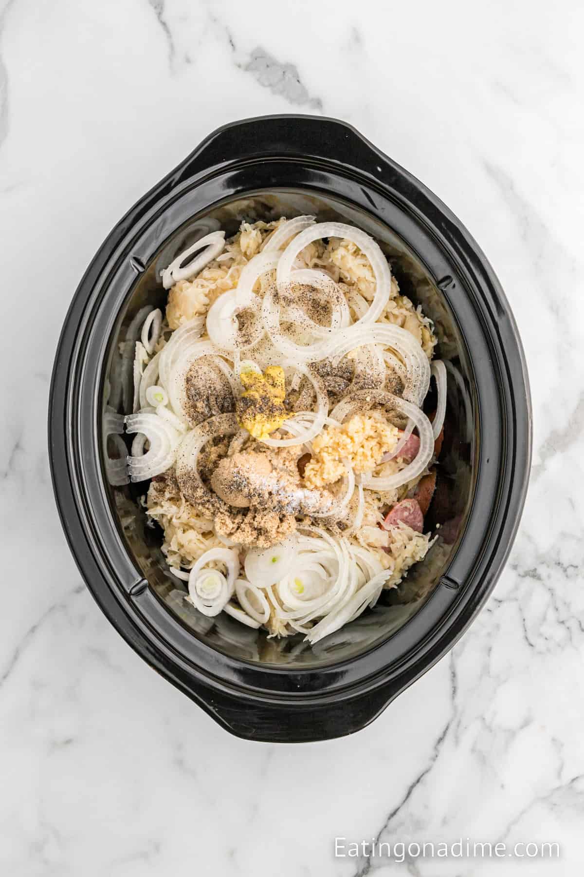 A black slow cooker on a marble countertop filled with sliced onions, seasoned chopped ingredients, and visible black pepper and minced garlic on top. The food looks uncooked. The website "Eatingonadime.com" is visible in the bottom right corner, featuring Slow Cooker Kielbasa and Sauerkraut recipes.