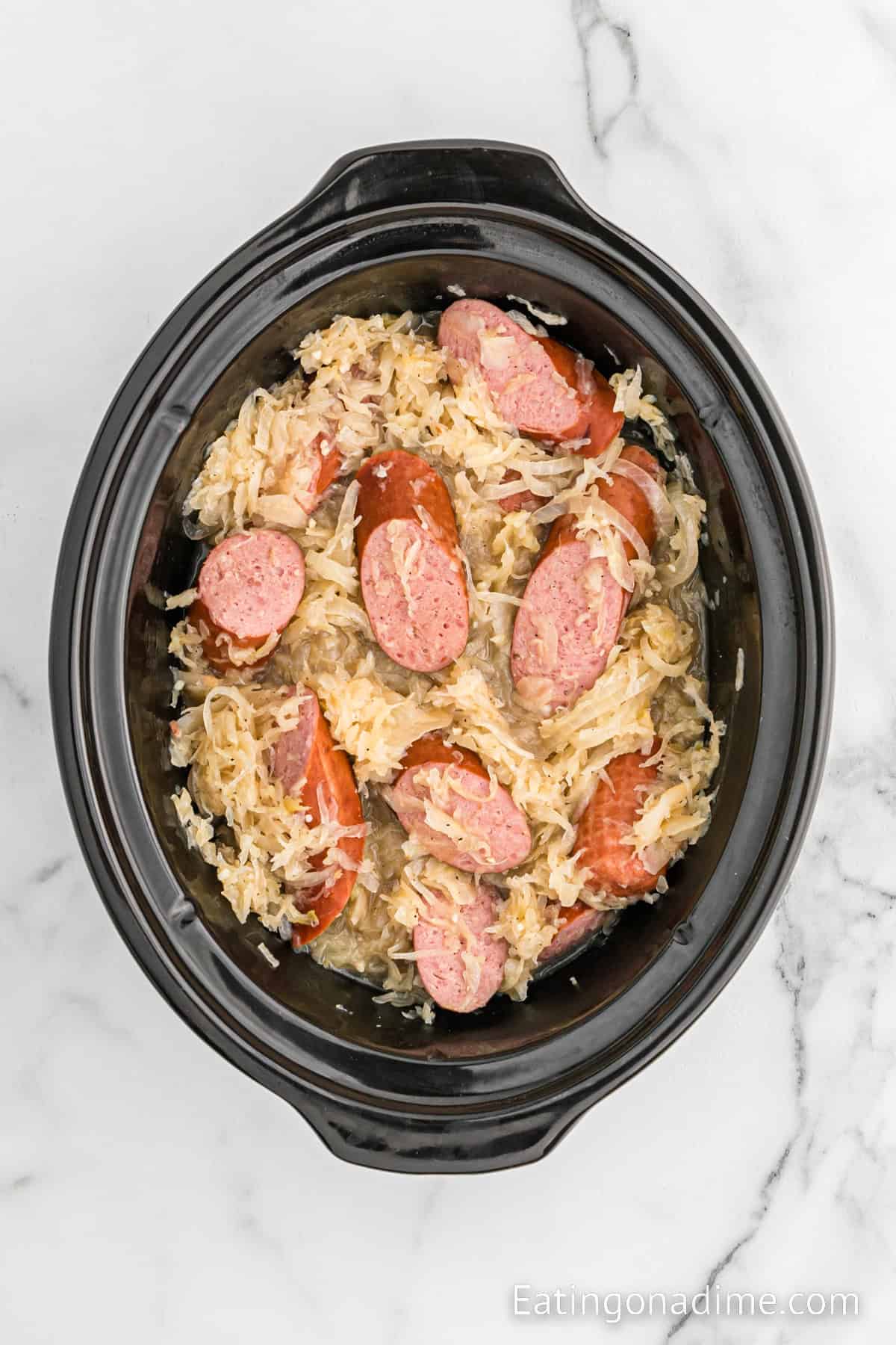 A black slow cooker filled with sliced kielbasa and shredded sauerkraut. The sausages are cooked and slightly browned, scattered among the pale sauerkraut. The slow cooker is placed on a white marble surface, creating the perfect Slow Cooker Kielbasa and Sauerkraut dish.