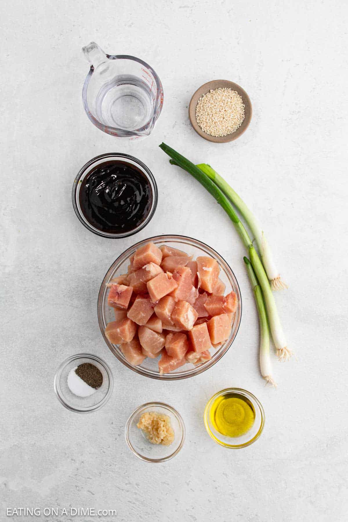 Ingredients for a Hoisin Chicken recipe are arranged on a white surface, featuring a bowl of cubed raw chicken, green onions, minced garlic, olive oil, salt, pepper, soy sauce, water in a measuring cup, and sesame seeds in a small bowl.